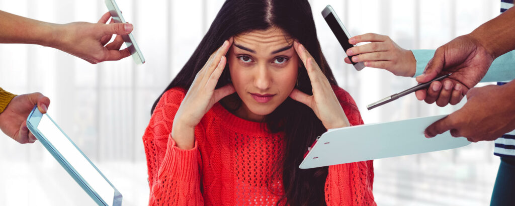 Jeune femme qui se tient le visage entre les mains et qui se dit "je n'arrive plus à travailler". Tout autour d'elle il y a des mains qui lui tendent des téléphones, des stylos, des tablettes.