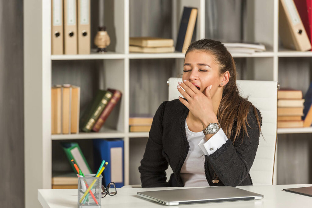 Femme assise à un bureau avec un ordinateur portable fermé devant elle. Elle baille, a les yeux fermé et une main devant sa bouche. Elle s'ennuie au travail.