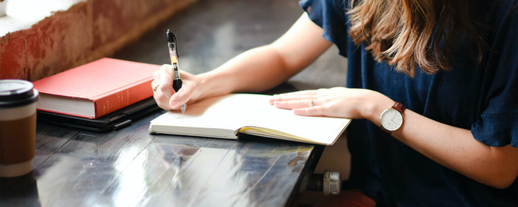 Une femme prend des notes dans un cahier. Elle est assise à une table.