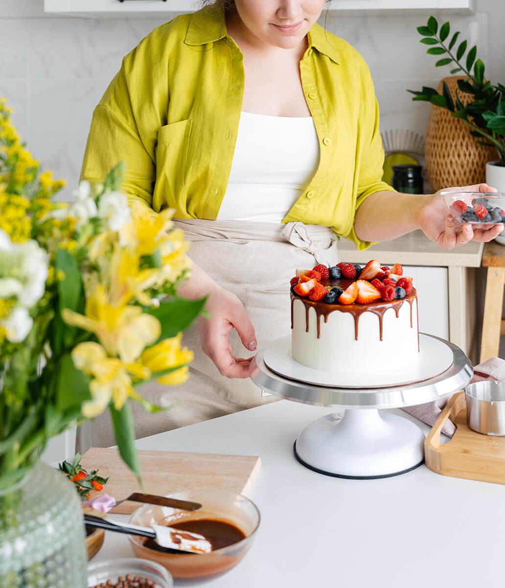 Une pâtissière est en train de poser des fruits sur un gâteau blanc couvert de chocolat.