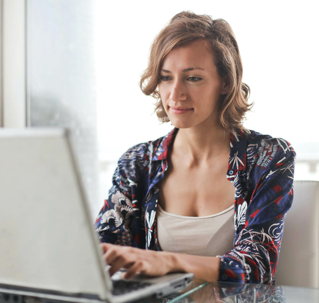 Une jeune femme assise à une table avec un ordinateur portable face à elle. Elle travaille sur le questionnaire enquête métier.