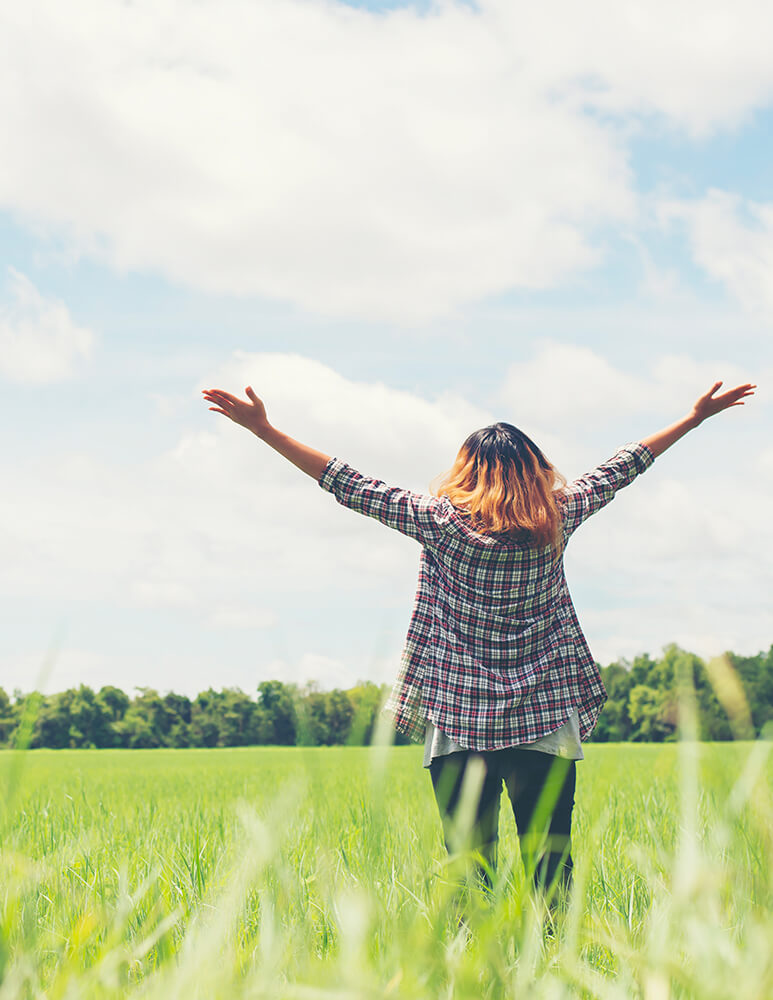 Jeune femme de dos dans un champ, elle lève les bras vers le ciel, elle semble heureuse et libérée d'avoir quitter l'enseignement.