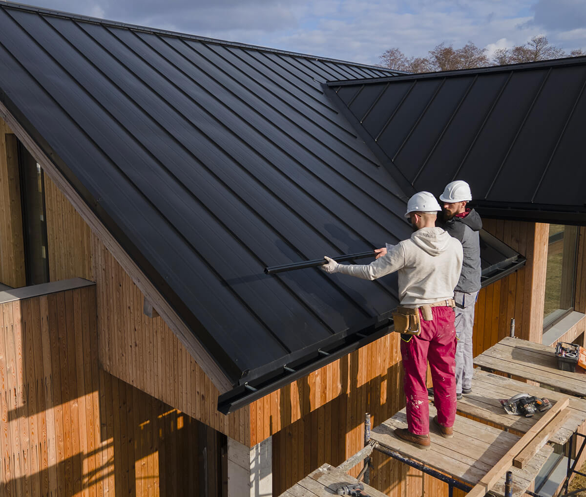 Deux couvreurs portant des casques de protection travaillent sur le toit d'une maison en bois. Ils sont en train d'installer ou de vérifier des panneaux métalliques noirs. L'un d'eux tient une grande barre métallique, tandis que l'autre ajuste les panneaux. Ils se tiennent sur un échafaudage en bois.