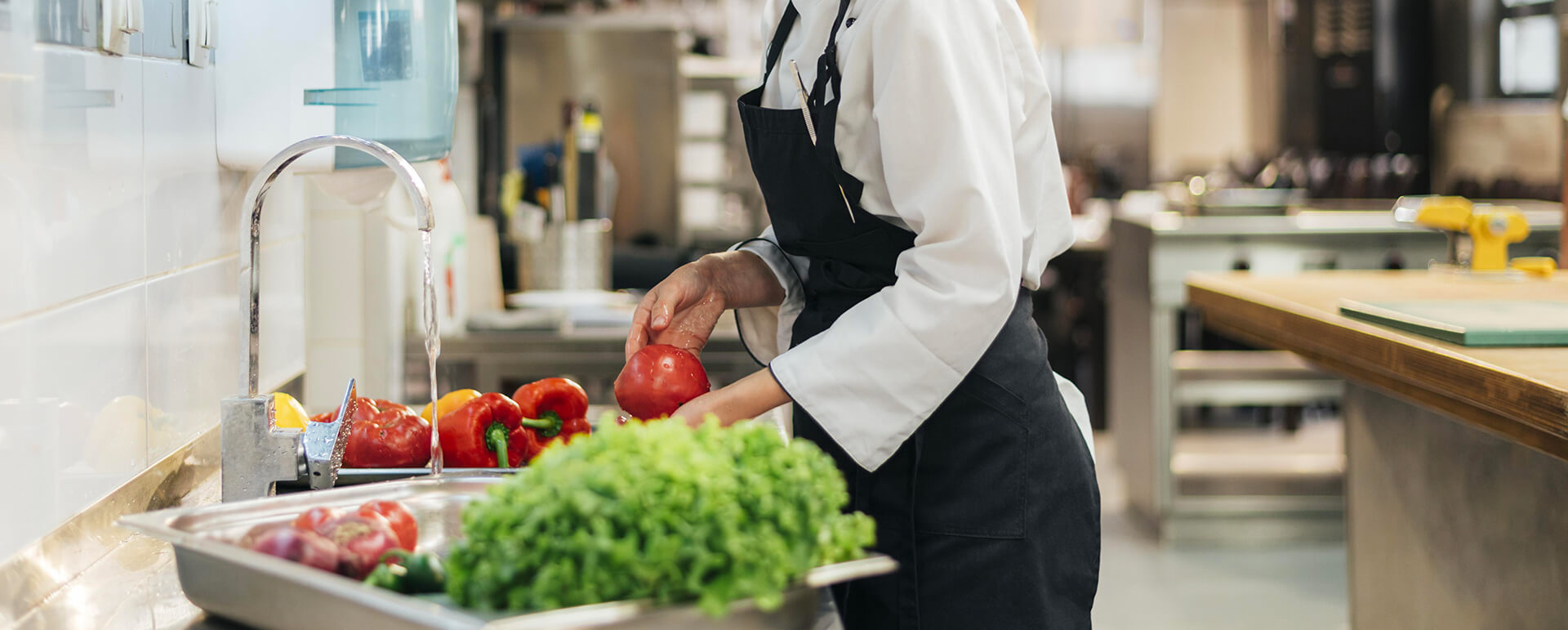 Un cuisinier en uniforme blanc et tablier noir lave des poivrons rouges sous un robinet d'eau dans une cuisine professionnelle. Sur le plan de travail en inox à côté, on voit d'autres poivrons et des légumes verts prêts à être préparés. L'environnement est propre et bien équipé, typique d'une cuisine de restaurant.