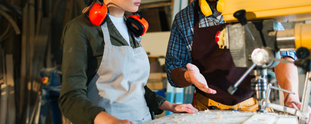 Deux personnes travaillent dans un atelier de menuiserie. La femme, en apprentissage après la troisième, portant un tablier gris et des protecteurs auditifs orange autour du cou, écoute attentivement l'homme à côté d'elle. L'homme, vêtu d'une chemise à carreaux bleue et d'un tablier marron, explique quelque chose en utilisant des gestes de la main près d'une machine à bois. Des outils et des matériaux de menuiserie sont visibles en arrière-plan, suggérant un environnement de travail manuel et technique.