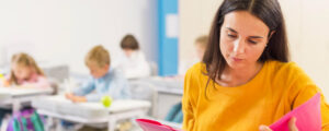 Une professeur des écoles habillée avec un pull jaune regarde des documents expliquant comment faire un bilan de compétences dans l'éducation nationale.