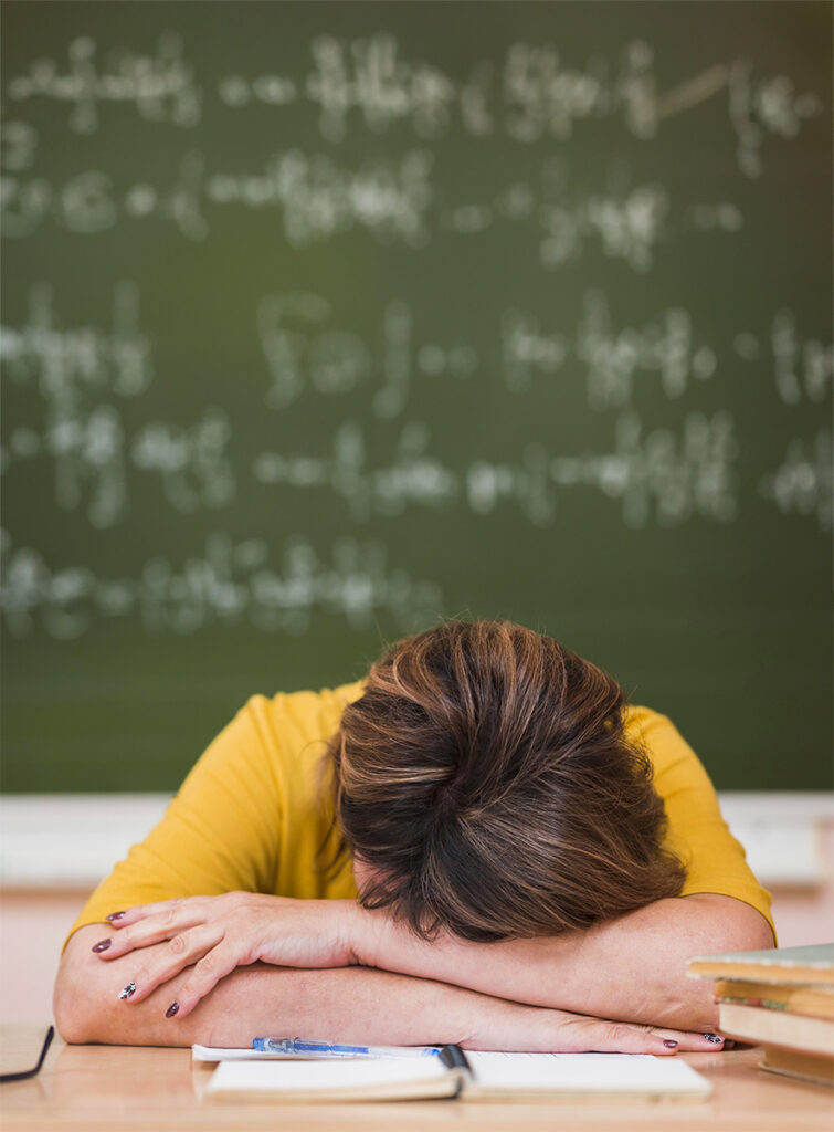 Une professeur a les bras croisés sur son bureau, sa tête repose dessus, on ne voit pas son visage. Elle semble épuisée et songe à quitter l'éducation nationale.