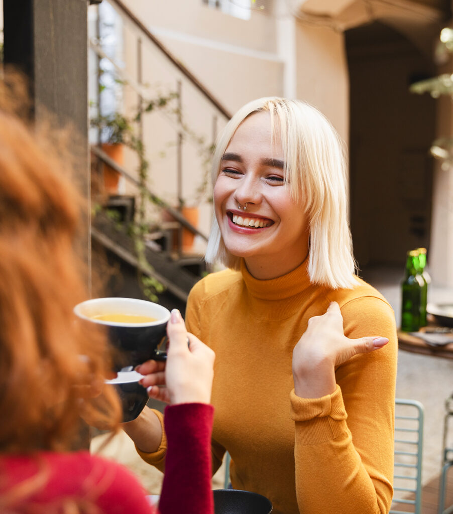 Une jeune femme blonde qui sourit fait face à une amie, de dos. Elles ont chacune une tasse dans la main.