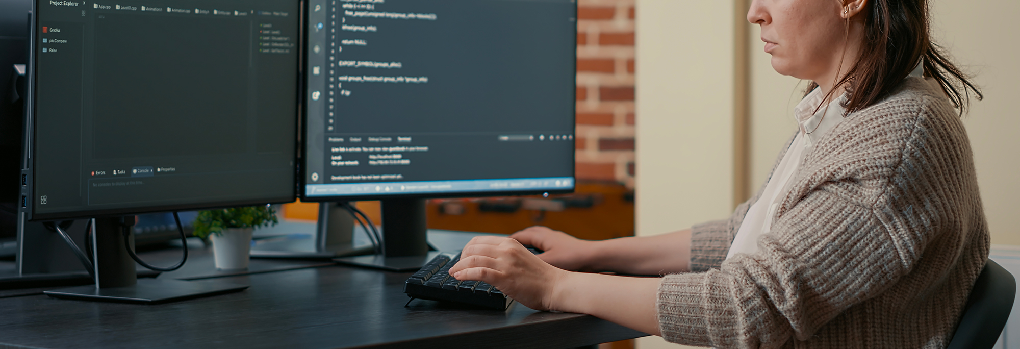 Une femme est assis à un bureau et fait face à deux écrans d'ordinateur sur lequel on voit des lignes de code informatique.