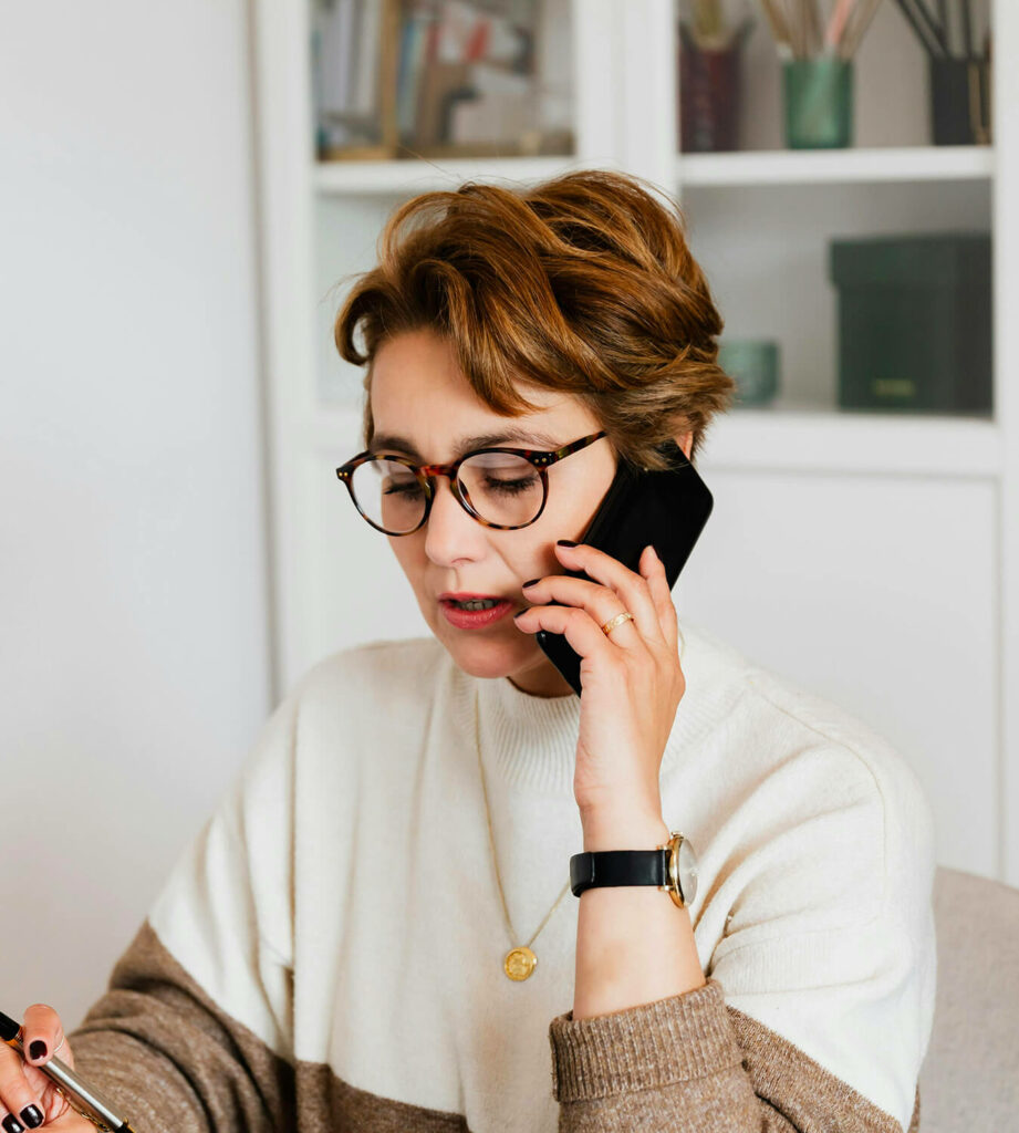 Une femme aux cheveux courts et qui porte des lunettes est au téléphone. Elle est à la recherche du meilleur bilan de compétences.